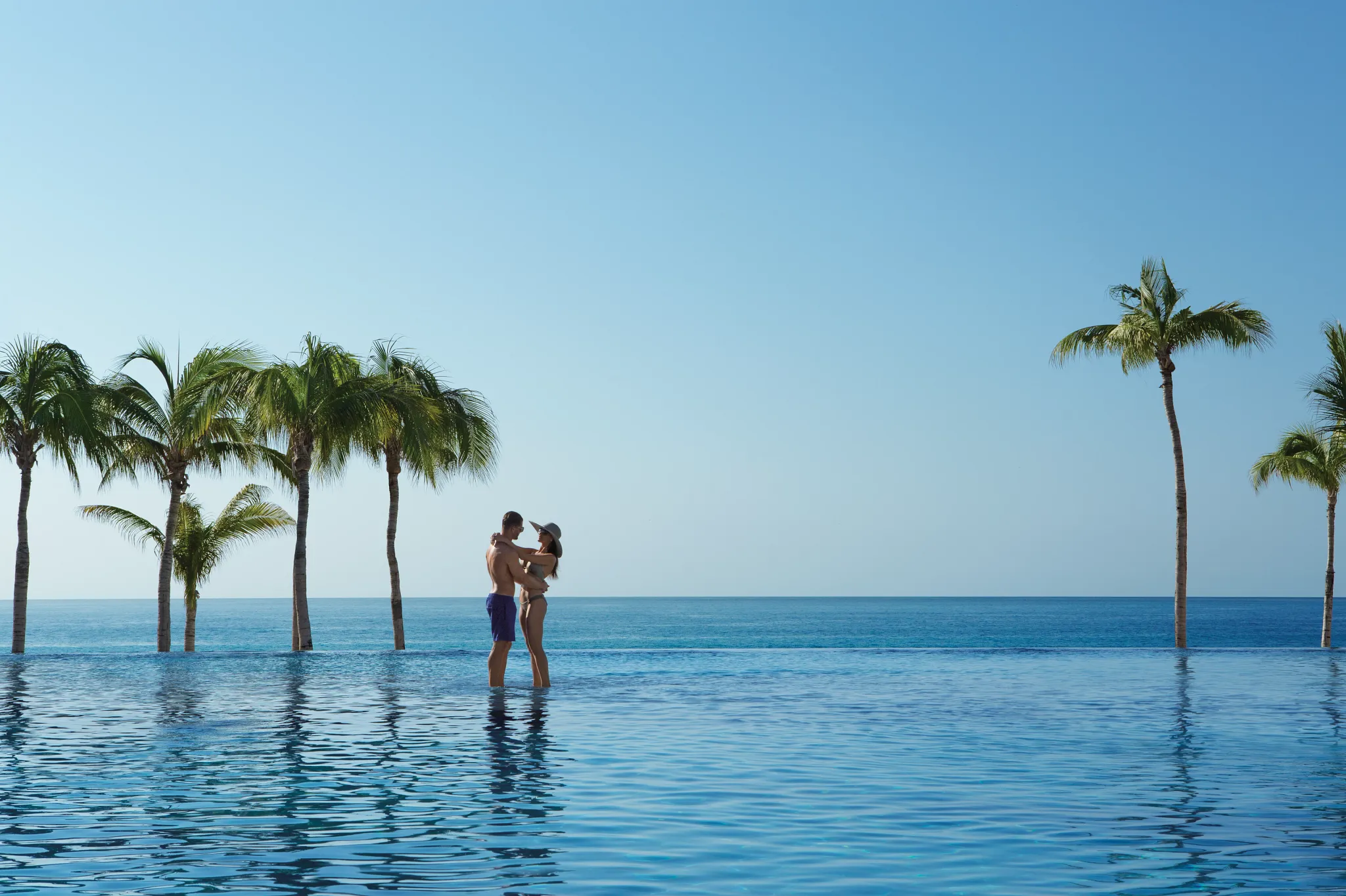 Couple embracing in an infinity pool framed by palm trees and ocean views at Dreams Los Cabos Suites Golf Resort & Spa.