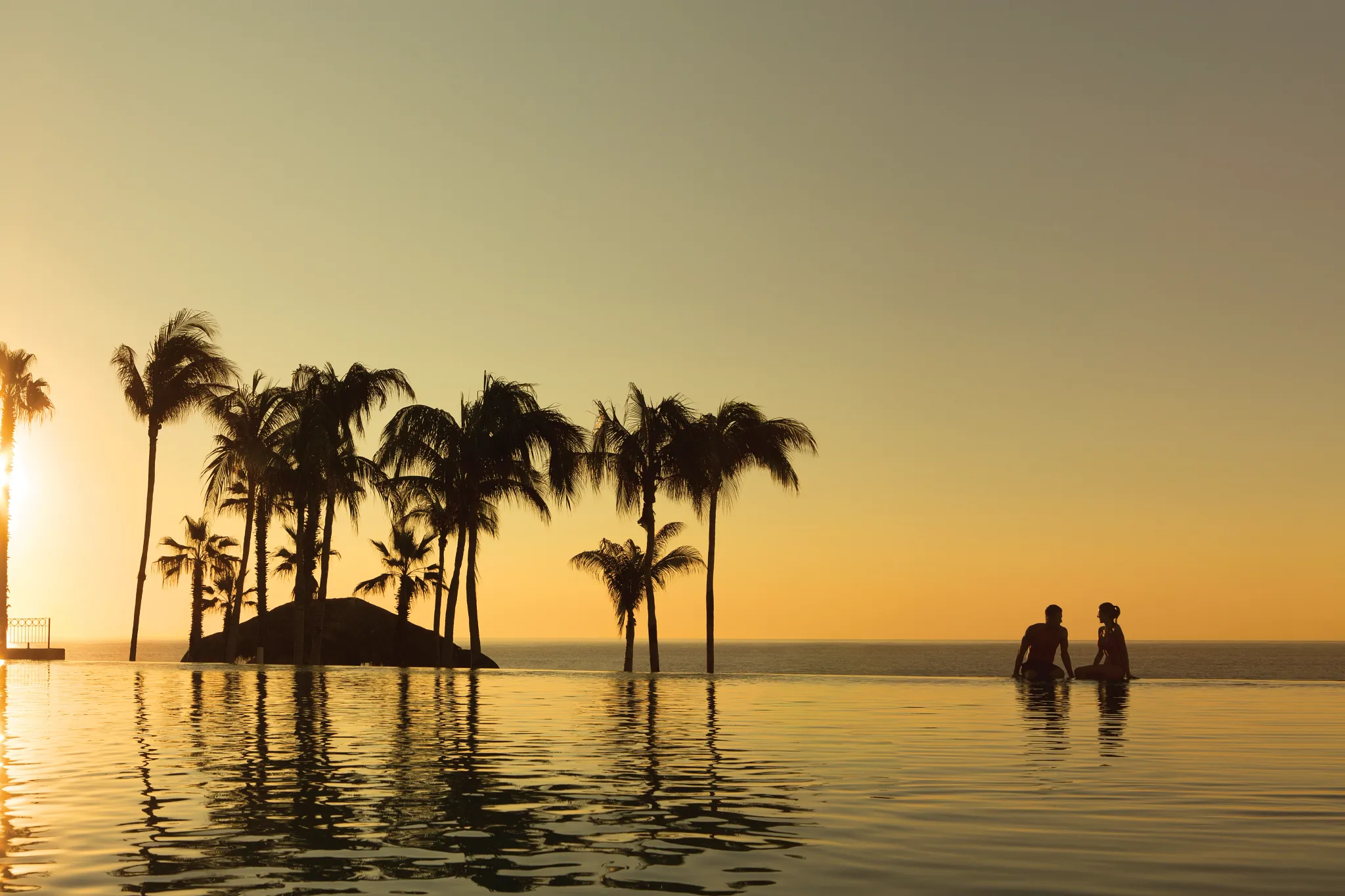 Couple sitting at the infinity pool during sunset overlooking the ocean at Dreams Los Cabos Suites Golf Resort & Spa.