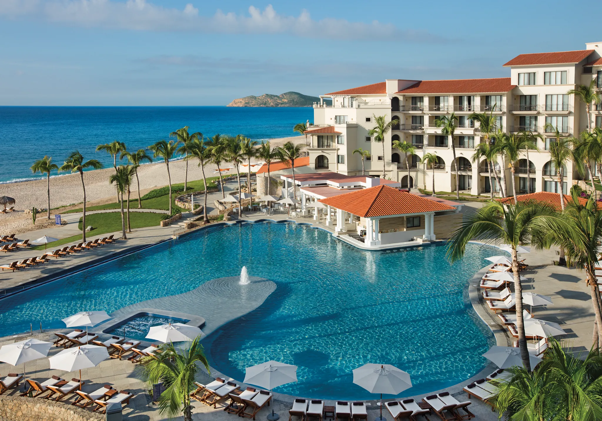Panoramic view of the oceanfront main pool surrounded by palm trees at Dreams Los Cabos Suites Golf Resort & Spa.