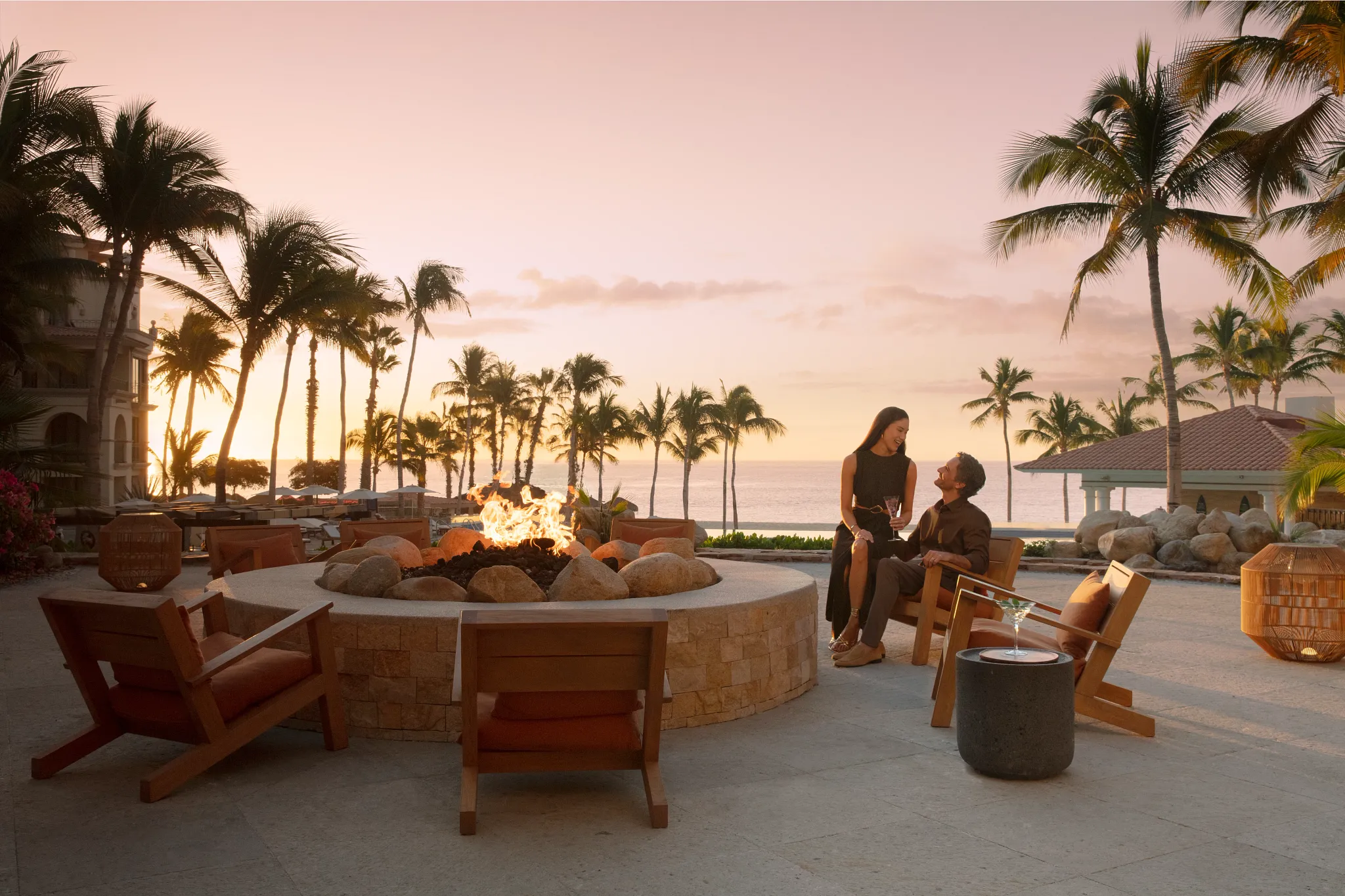 Couple enjoying sunset drinks beside an oceanfront fire pit at Dreams Los Cabos Suites Golf Resort & Spa.