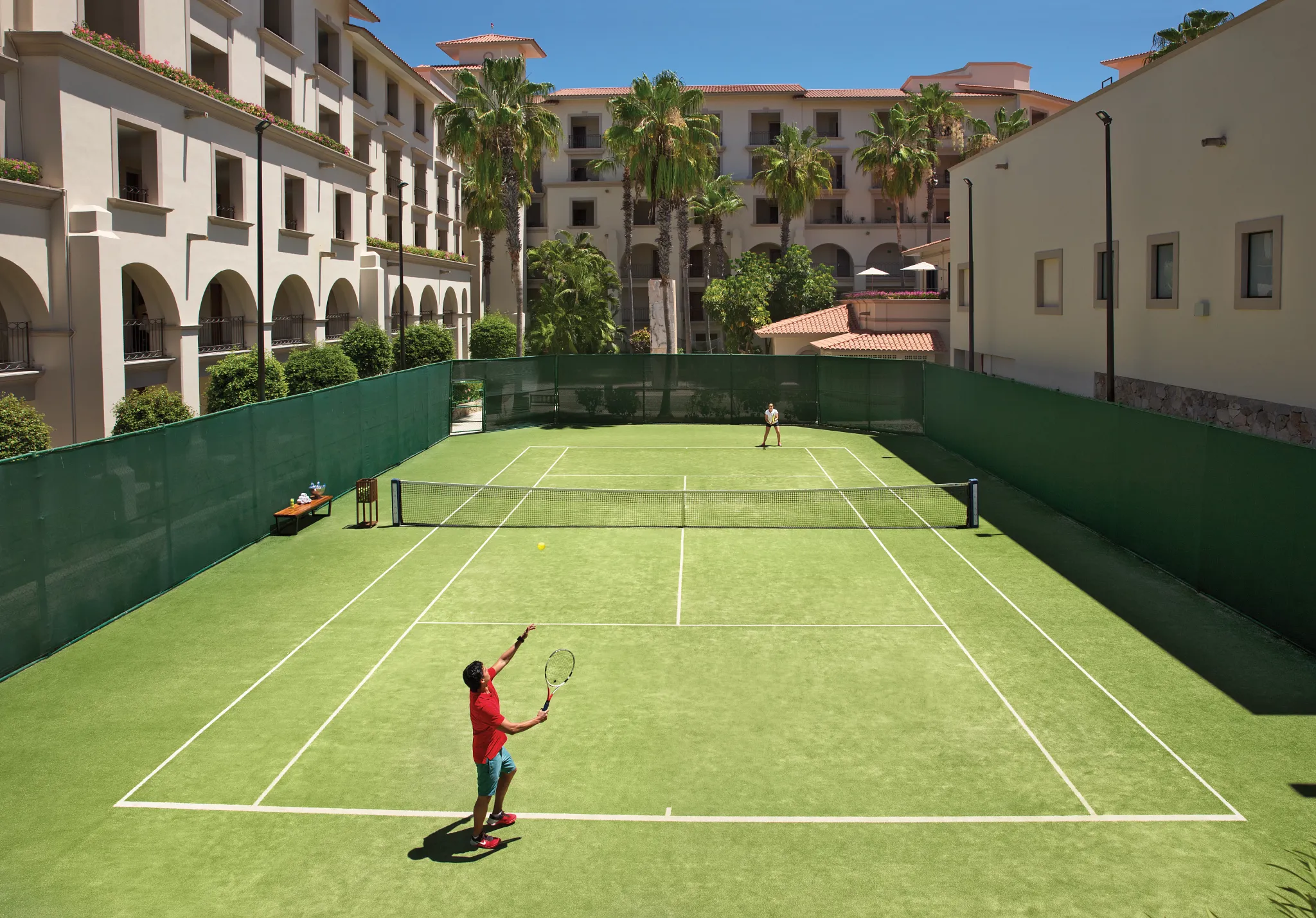 Guests playing tennis on the outdoor resort tennis court surrounded by palm trees at Dreams Los Cabos Suites Golf Resort & Spa.